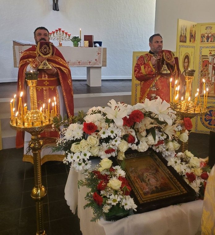 Ікона на столі серед білих і червоних квітів, обабіч високі свічники, позаду духовенство в червоних ризах