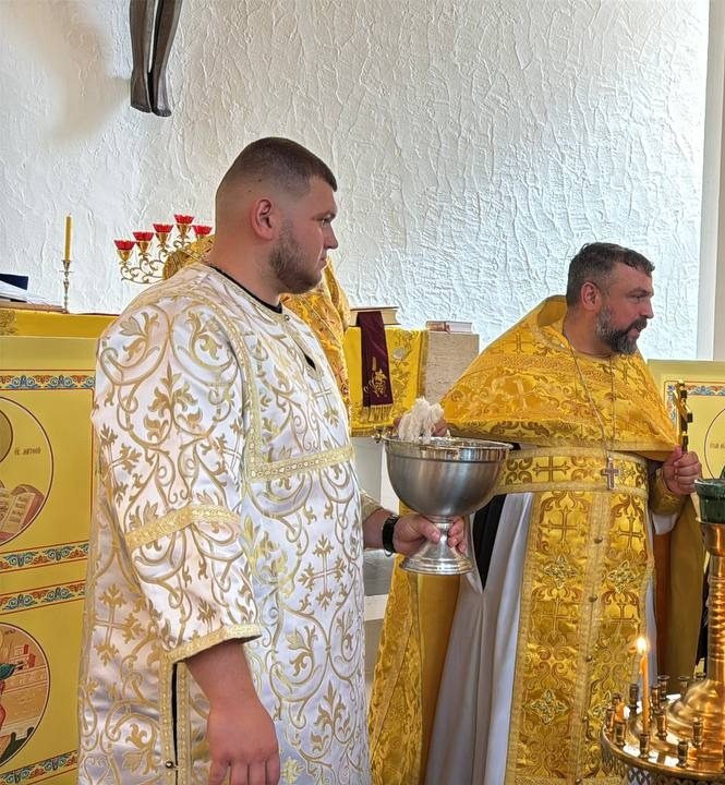 Прислужник у білому стихарі тримає срібну посудину зі свяченою водою й кропилом, поруч священник у золотих ризах
